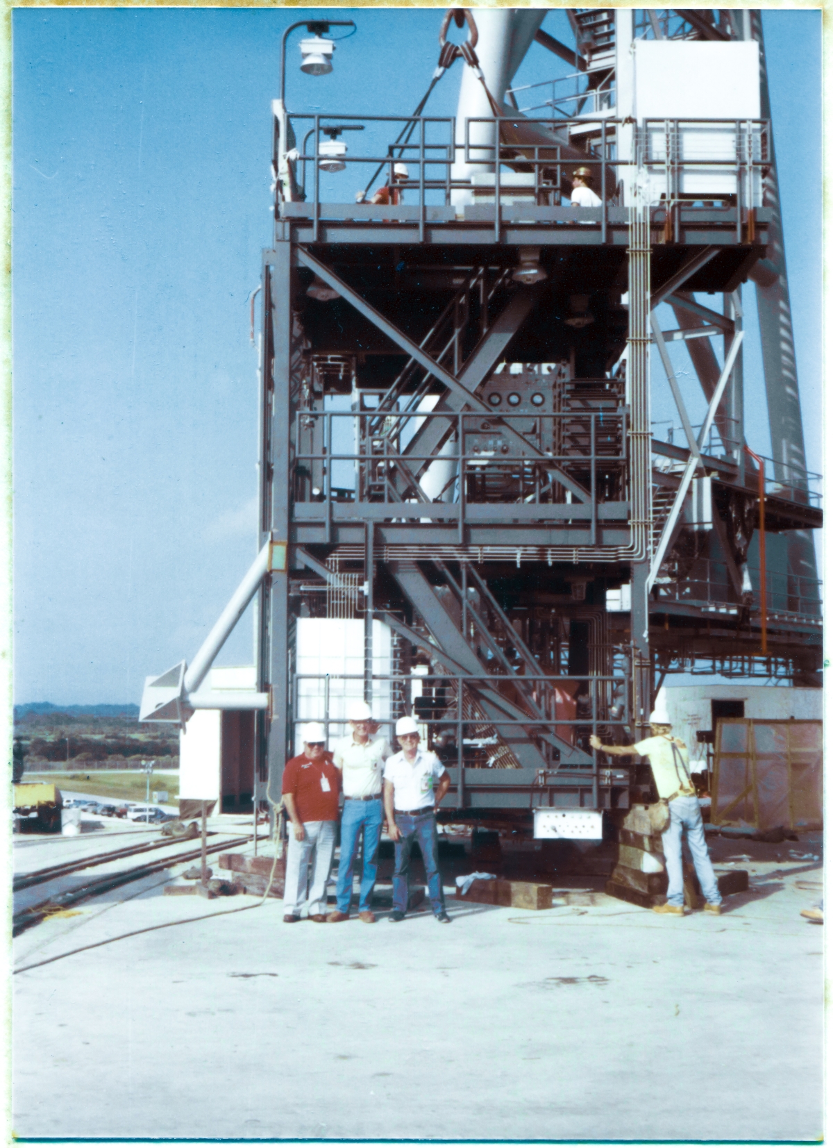 Image 086. The OMBUU. The Orbiter Mid-Body Umbilical Unit, sitting on the Pad Deck at Space Shuttle Launch Complex 39-B, moments prior to being lifted into place, high above, flush against the face of the RSS. Standing in front of it, shoulder-to-shoulder like the Three Musketeers, left-to-right, Howard Baxter, Wade Ivey, and Hank Morgan smile widely, thoroughly enjoying a momentary breather from the high-stress demands of their intimate involvement with yet another dangerous and high-stakes operation which is just about to get underway, provided by James MacLaren making a silly remark to them an instant before hitting the shutter release on his camera. To their right, back to camera, Rink Chiles' brother Rayburn, another Union Ironworker from Local 808, leans against the corner of the OMBUU, which is still sitting on temporary support cribbing, getting ready to feel it come alive and go into weightless motion, and steadying himself against any drift which the OMBUU might take once it goes fully into suspension and breaks free of the ground. Up on top of the OMBUU, another pair of, unknown alas, Union Ironworkers prepare to take the stairs down to the lowest level and get off of it, following fine adjustment and close inspection of the four-legged lifting sling which will be what carries it aloft, and which is already in tension. The OMBUU is a ferociously-complex and potentially explosive piece of equipment, and its main job is to pump Liquid Hydrogen and Liquid Oxygen at unimaginably-cold temperatures into the Dewars beneath the Orbiter's Cargo Bay which will store them as consumables for the entire duration the Shuttle remains on orbit. Photo by James MacLaren.